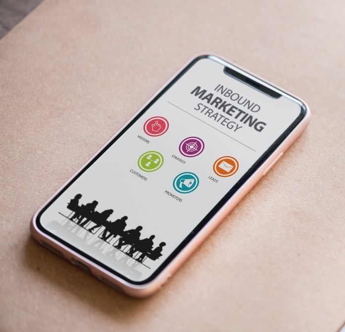 A imagem mostra a tela de um celular contendo a frase Inbound Marketing Strategy (Estratégia de Inbound Marketing)  e cinco ícones coloridos, cada um representando um item - visitors (visitantes), strategy (estratégia), leads, customers (clientes) e promoters (promotores). Abaixo, aparece uma silhueta de um grupo em volta de uma mesa de reuniões.